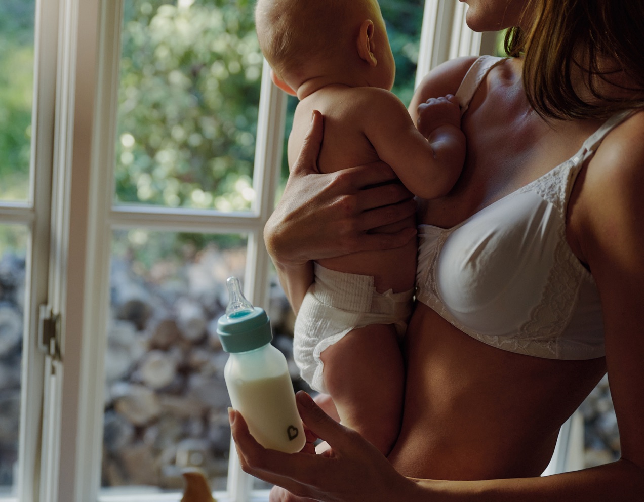 A mother holding a baby and a bottle beside a bright window