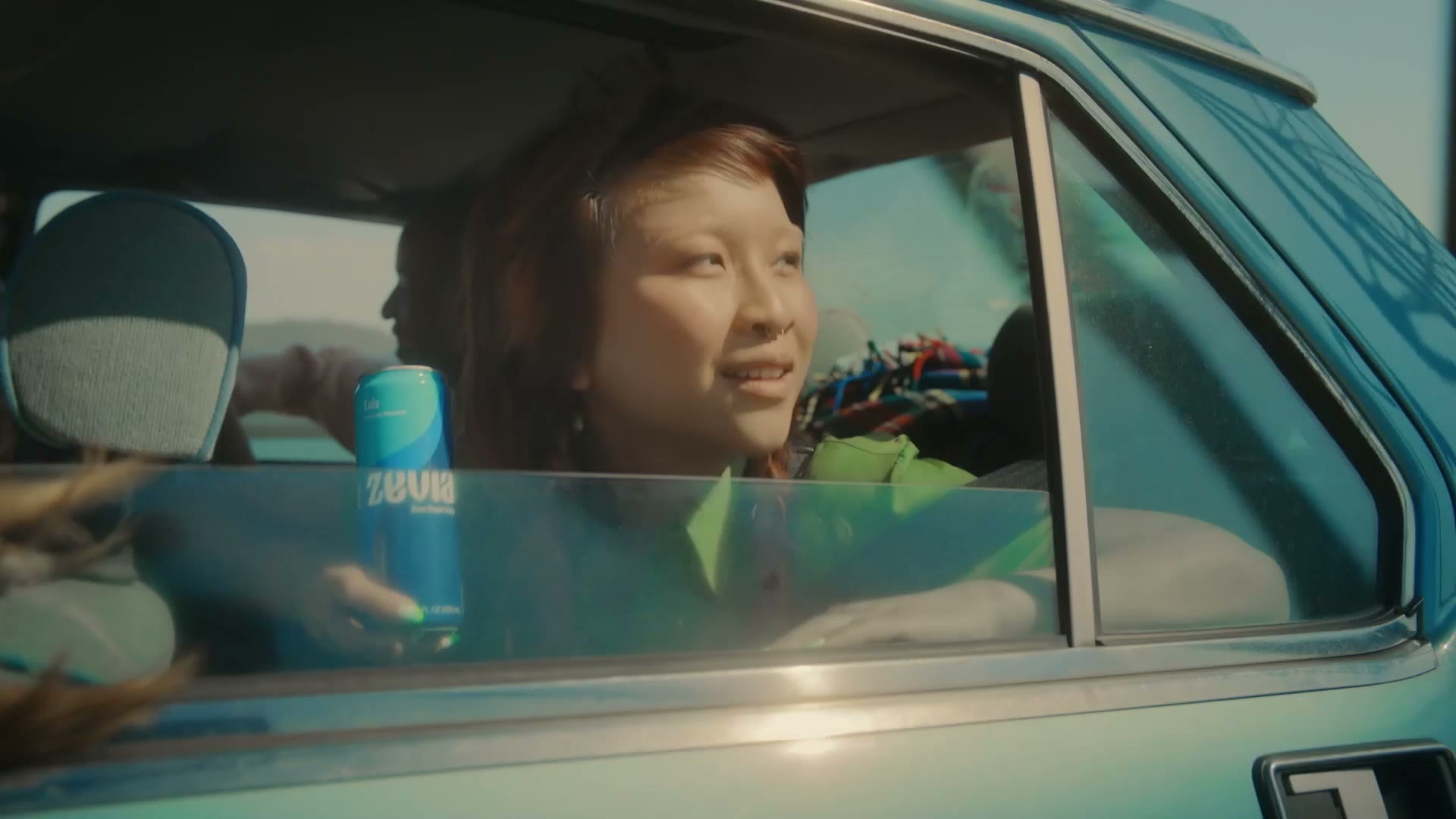 A woman smiling out of a blue car window, holding a Zevia can